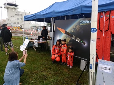 宇宙服を着た親子がロケットが描かれたパネルの前で記念撮影している写真