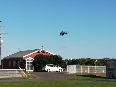 青空の中、機体を傾けて飛行しているヘリコプターが、赤い平屋の建物とともに収められた写真