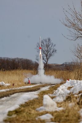 曇り空のなか雪の残る地面の奥でロケットが打ち上げられている様子が収められた写真