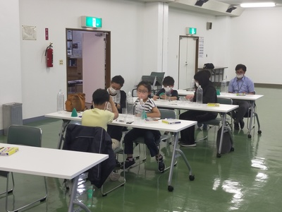 中会議室内に設けられた机の上でペットボトルロケット製作に臨む団員児童たちとその保護者たちの写真