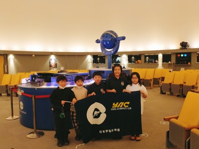 釧路こども遊学館館内のプラネタリウム投影機の前で、YAC団旗を掲げつつ記念撮影に応じる団員児童たち5名の集合写真