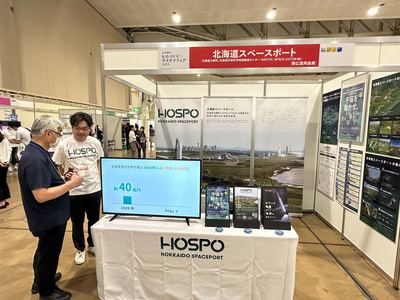 北海道スペースポートのブースの前面にHOSPOと書かれたディスプレイとパンフレットの置かれた机があり、壁にポスターが貼られている様子の写真