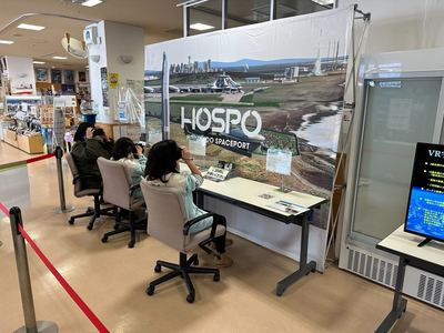HOSPOのパネルが配置されたコーナーで椅子に座ってVRゴーグルをつけ、VR宇宙旅行体験をする3人の写真