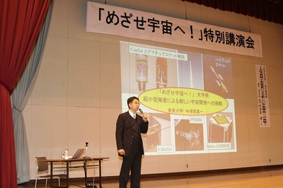 体育館のステージ上でスライドショー形式にて進行する、「めざせ宇宙へ!」特別講演会の講演風景の写真