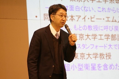 体育館のステージ上でマイクを片手に講演を行う中須賀真一教授の写真