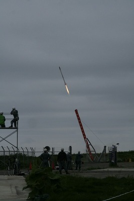 曇り空のなか、海上に向けて斜めの発射台から打ち上げられているロケットの写真