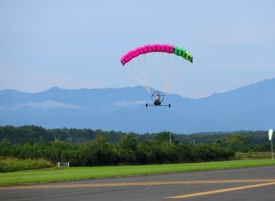 青空と山をバックにした、桃色と緑色の翼を広げて滑走路の上を飛行しているパラグライダ型UAVの写真
