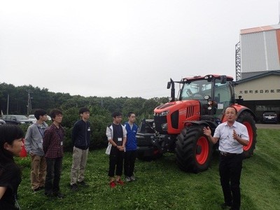 多目的航空公園内の格納庫付近にて、農業用トラクターを前に講義を受ける参加者たちの写真