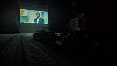 暗い夜中、除雪された路面に車が止まっており、格納庫のシャッターに映画が投影されている様子の写真