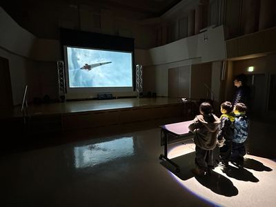 発射ボタンが置かれた中央の机の前に立つ参加者と、ステージ上に設けられたスクリーンに投影されたロケットの映像の写真