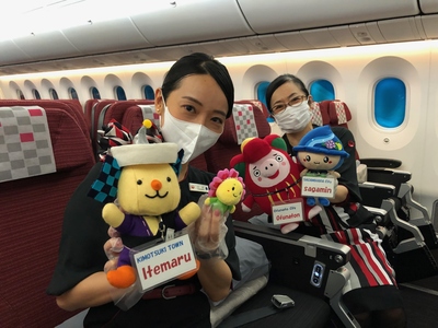 旅客機キャビンにて、各銀河連邦共和国のマスコットキャラクターのぬいぐるみ4種(左から肝付町のいて丸、大樹町のコスピー、大船渡市のおおふなトン、相模原市のさがみん)を手に持って撮影に応じるJALのCA2名の写真