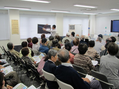 飛行管制棟内の一室にて、着席して職員からの解説を聞く幕別桂寿会の訪問団の写真