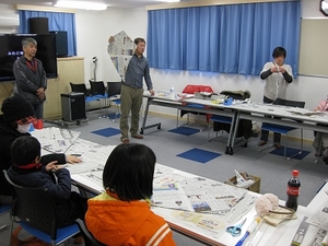SORA施設内にて、参加した児童たちへ新聞紙製の凧の製作についてレクチャーを行う講師の写真