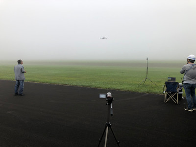 濃霧の中で実施された、ドローンの飛行試験風景の写真。写真中央やや右上に低空飛行中のドローン、写真手前に記録用カメラ、写真両端に操縦・観測を行うスタッフがそれぞれ確認できる
