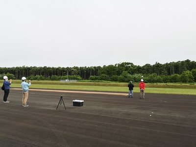 晴れた滑走路上で実施された、ドローンの飛行試験風景の写真。写真左端に観測するスタッフ、右側奥に操縦するスタッフの姿が確認できる