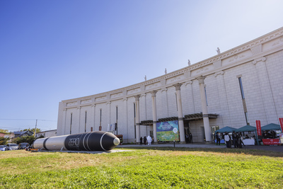芝生にロケットの模型が置かれ、その奥にベルクラシック帯広の白い建物があり、入り口前にテントが並んでいる様子の写真