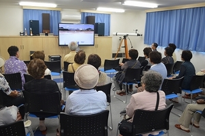 SORAの展示室2にて着席して資料映像を鑑賞する、帯広市わかば会・緑わかば会の見学団の写真