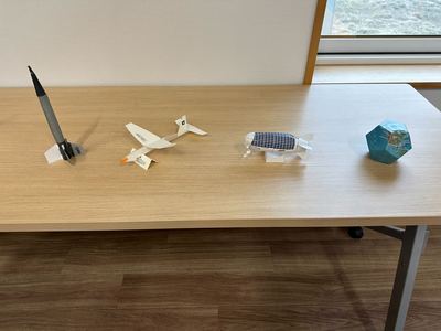 左から先端部と後ろ側が黒く胴体が灰色のペンシルロケット、尾翼まで含めた飛行機の形をしたホワイトウィングス、上側が紺色の飛行船の形をした成層圏プラットフォーム、正十二面体の形の地球儀貯金箱の4種類のペーパークラフトが並んでいる様子の写真