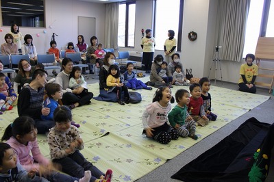視聴覚室で開催されたクリスマスのつどいで、シートの上や椅子に座り、たんぽぽ会員の方の出し物を楽しむ子供たちの写真