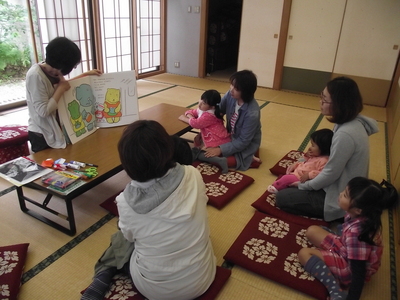 児童室で絵本の読み聞かせをするボランティアさんと楽しそうに聞き入る子どもたちと保護者の方々の写真