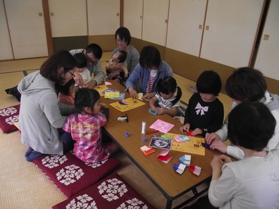 机の周りに囲むように座り、折り紙を楽しむボランティアスタッフと子供たちと保護者の方々の写真