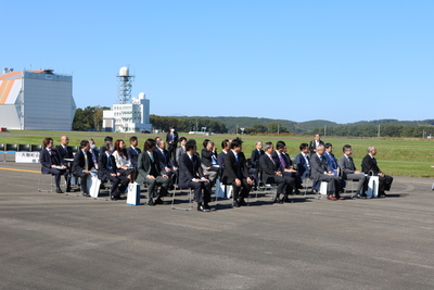 晴天の滑走路上に設けられた席に着座して式典に臨む各参加企業代表たちを、式典会場右側前方よりおさめた写真