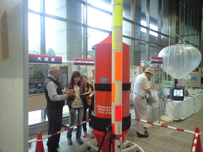 ガラス張りのエリアに展示されたロケットの前で来場者に解説をするスタッフの写真