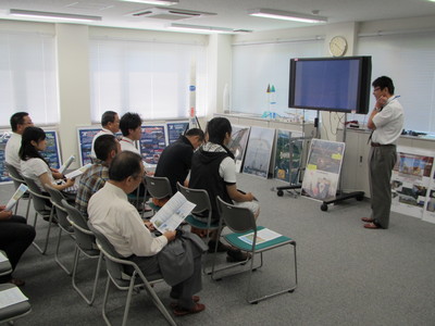 飛行管制棟内の一室にて、各々着席して資料を手看しつつ職員からの解説を聞く、十勝三菱自動車販売株式会社の視察団第1陣の写真