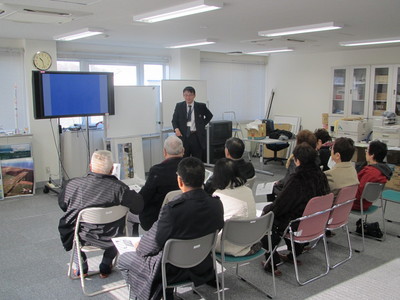 飛行管制棟内の一室にて着席して職員からの解説を聞く、元・尾田小中学校卒業生の訪問団の写真