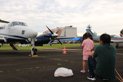 駐機した双発飛行機を正面から眺める女児とその保護者の写真