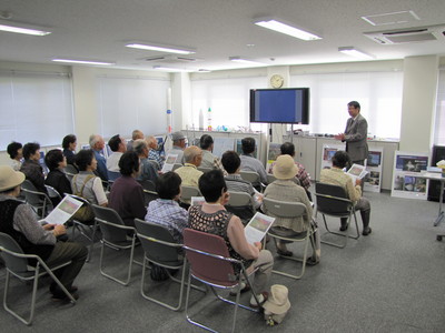 飛行管制棟内の一室にて着席して職員からの解説を聞く、中伏古老人会の見学団の写真