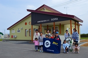 SORAの建物前にて記念撮影に応じるYAC大樹分団員たちの集合写真。写真中央右側には団員の児童2,3名がYACの団旗を掲げている様子が確認できる
