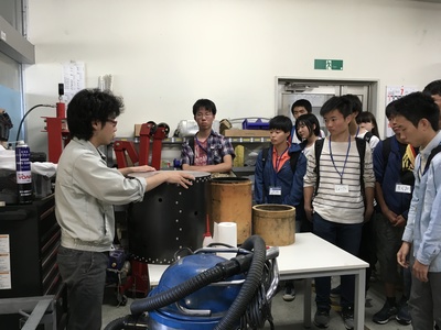 施設内の作業現場にて機材を前に職員からの解説を聞く高校生参加者たちの写真