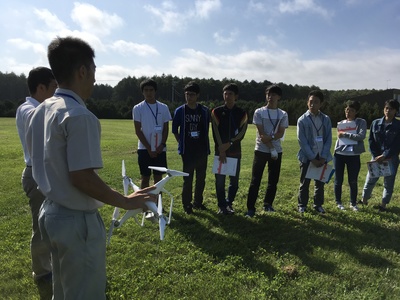 屋外の芝生の上で、小型ドローンを手にした講師より講義を受ける高校生参加者たちの写真