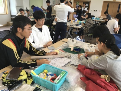 屋内の卓上にて、数人単位の班に分かれて各々モデルロケットの製作に臨む高校生参加者たちの写真