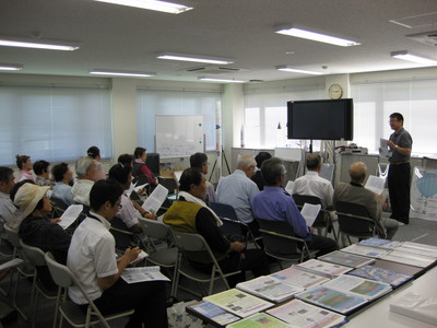 飛行管制棟内の一室にて、着席して職員からの解説を聞く帯広発明協会の訪問団の写真