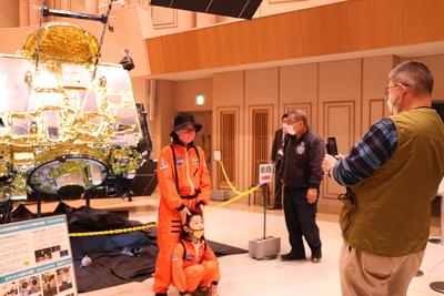 展示された「はやぶさ2」の実物大模型の前で、NASAの宇宙服姿で記念撮影を行う来場者親子の写真