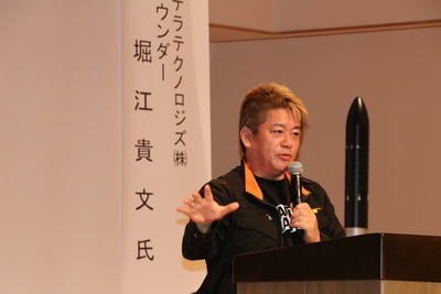 ロケットの模型の隣でマイクを持ち講演をしている堀江貴文氏の写真