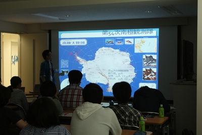 プロジェクタースクリーンを用いた座学の実施風景の写真。写真では気球を用いて大気採取を実施した、第54次南極観測隊に関する画面がスクリーンに投影されている様子が確認できる