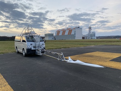 滑走路の端に停車した、オオワシ2号実験用ワゴン車の写真。車体には固定用器具が増設されており、その前方先端にオオワシ2号機縮小機体が設置されているのが確認できる