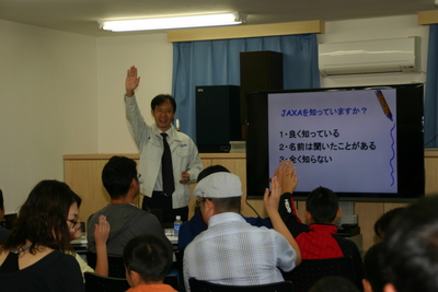 SORAの施設内における高橋氏の講演風景の写真。氏の隣に置かれたモニターの画面には「JAXAを知っていますか？」という問いのアンケート画面が表示されている