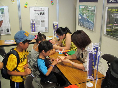 出展ブースで折り紙でのロケット製作を教わっている子供達の写真
