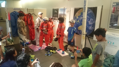 「2018サイエンスパーク」の出展スペース内にてNASAの宇宙服のレプリカの試着に臨む児童達の写真