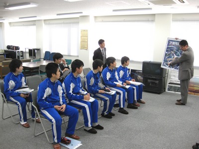 飛行管制棟内の一室にて、着席して職員からの解説を聞く豊似中学校の生徒たちの写真