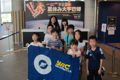 夏休み大宇宙博の会場入口付近にて記念撮影に応じる、研修旅行団の集合写真。写真中央手前には児童3名がYACの団旗を掲げている様子が確認できる