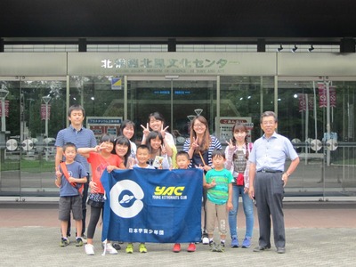 北網圏北見文化センターのエントランス前にて記念撮影に応じる研修旅行団の集合写真。中央最前列には団員の児童3名がYACの団旗を掲げている様子が確認できる